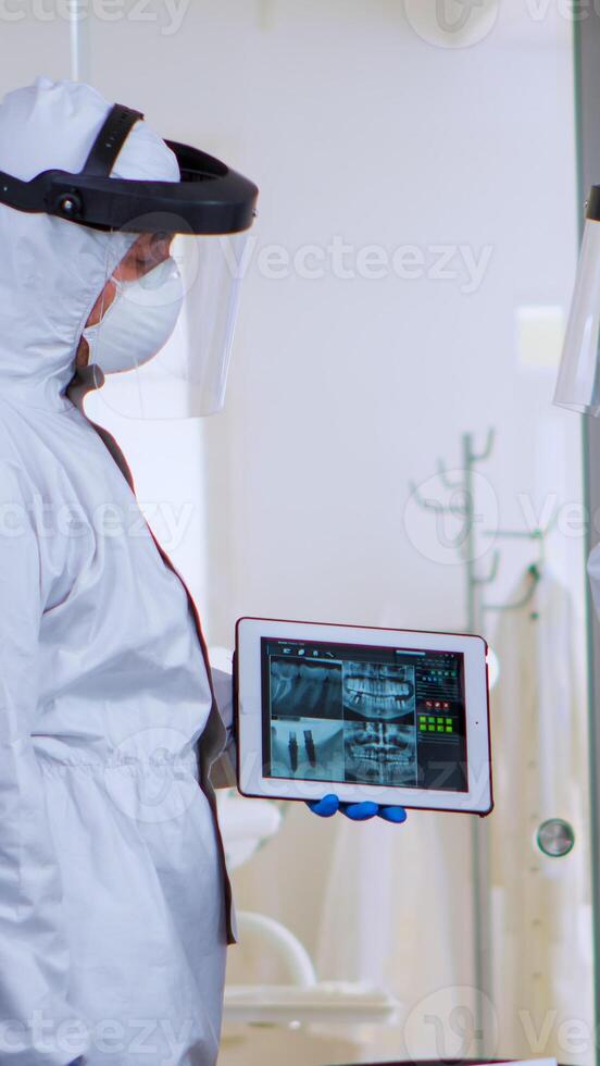 Orthodontics doctors with face shield and ppe suit discussing in reception about teeth digital x-ray using tablet during global pandemic. Concept of new normal dentist visit in coronavirus outbreak. photo