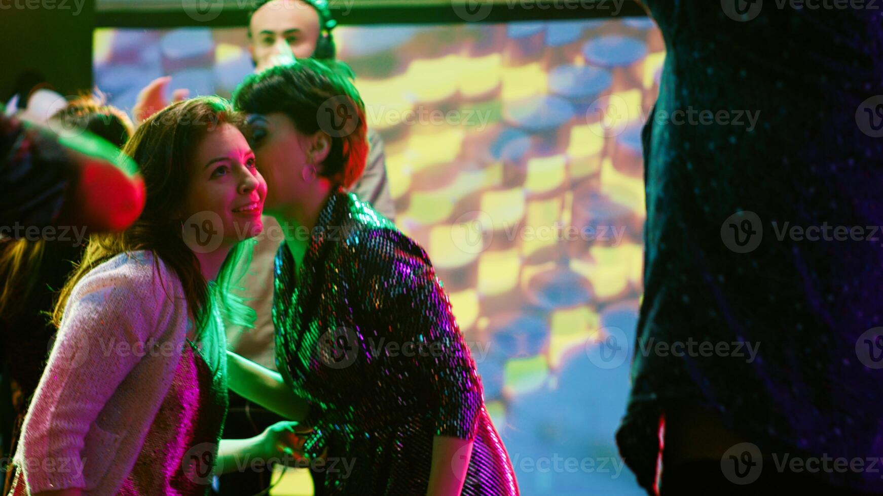 Happy girls chatting at nightclub, partying and trying to talk to each other while DJ mixes music on stage. Smiling women having fun on dance floor with multiple people, nightlife concept. photo