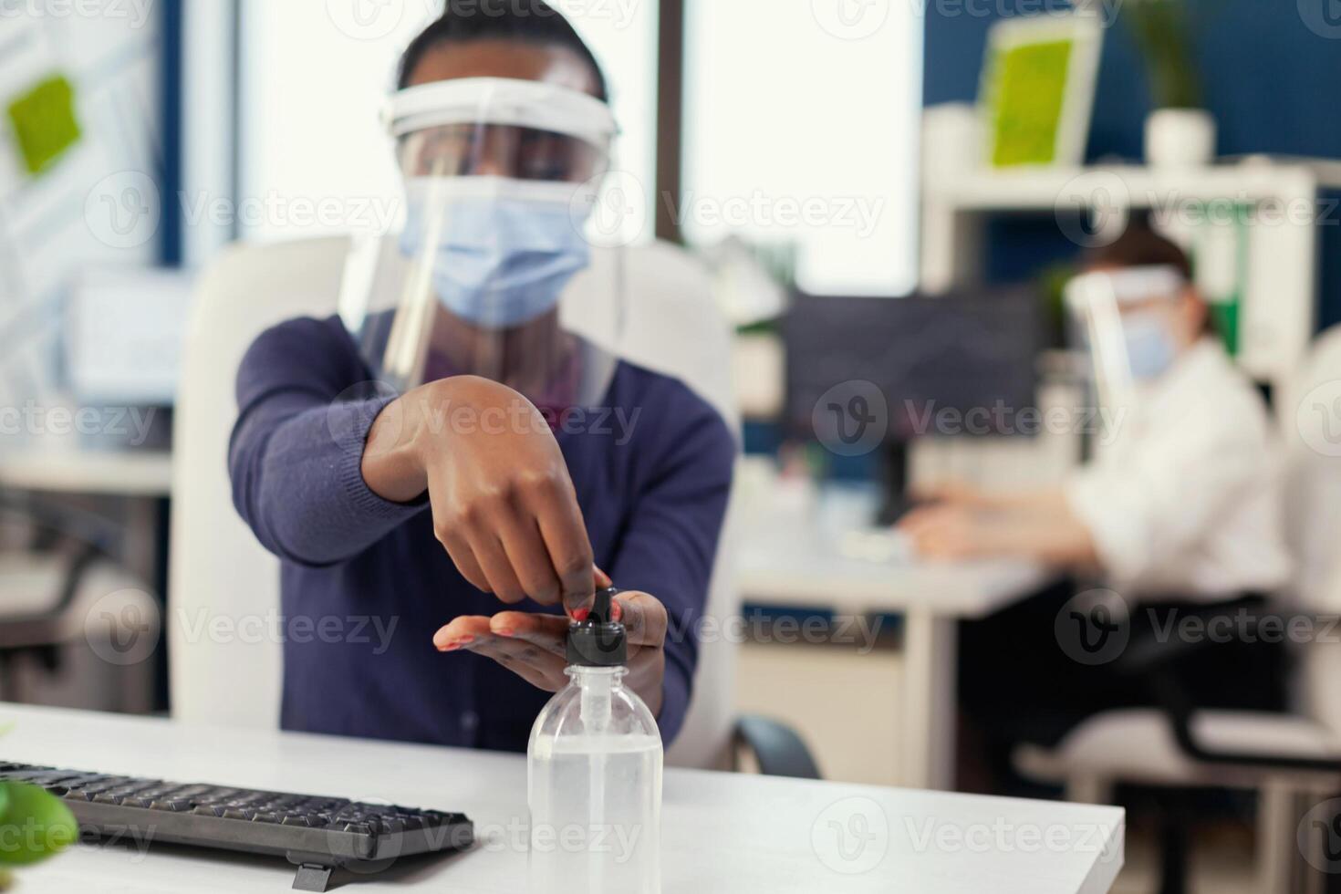 cerca arriba de africano mujer utilizando mano desinfectante durante global pandemia con coronavirus. mujer de negocios en nuevo normal lugar de trabajo desinfectar mientras colegas trabajando en antecedentes. foto