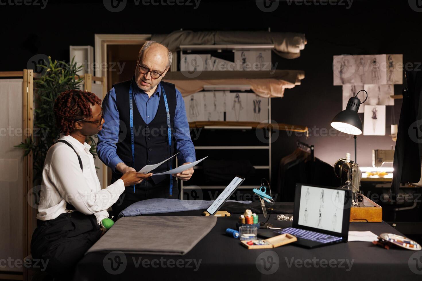 African american needleworker apprentice helping master couturier pick best sartorial outfit design for runway show collection. Tailor and assistant brainstorming ideas for creative bespoken attire photo