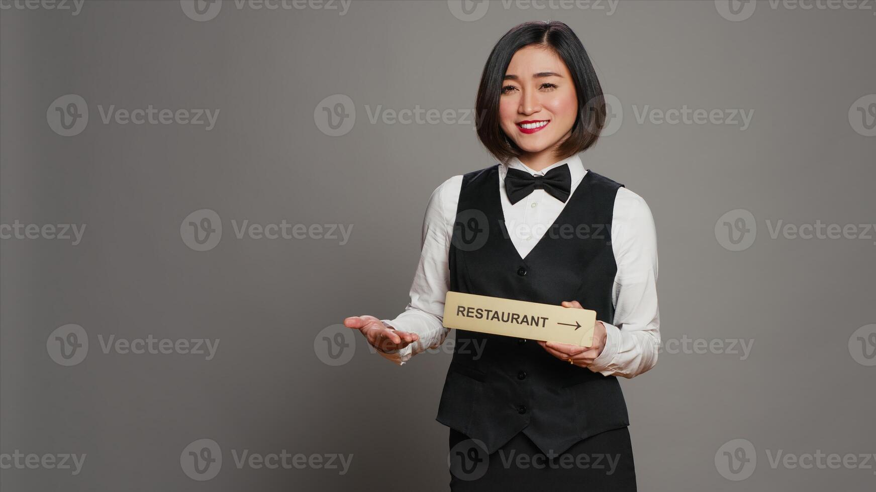 Asian hotel concierge holding restaurant sign to indicate direction, pointing towards dining area. Receptionist assisting clients to enjoy all amenities, stands over grey background. Camera A. photo