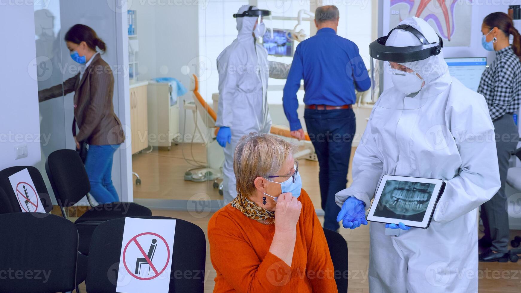 Dentist doctor with face shield pointing on tablet display explaining dental x-ray to senior patient during global pandemic. Nurse wearing protection suit, overall, mask and gloves, new normal photo