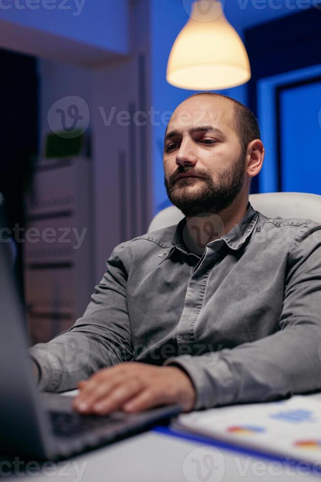 Serious young manager working on deadline doing overtime in empty office. Smart businessman sitting at his workplace in the course of late night hours doing his job. photo