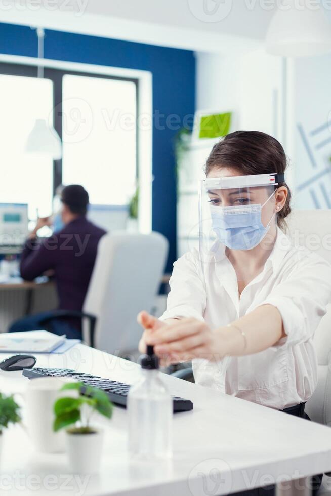 Office worker following safety precaution during global pandemic with coronavirus applying sanitizer. Businesswoman in new normal workplace disinfecting while colleagues working in background. photo