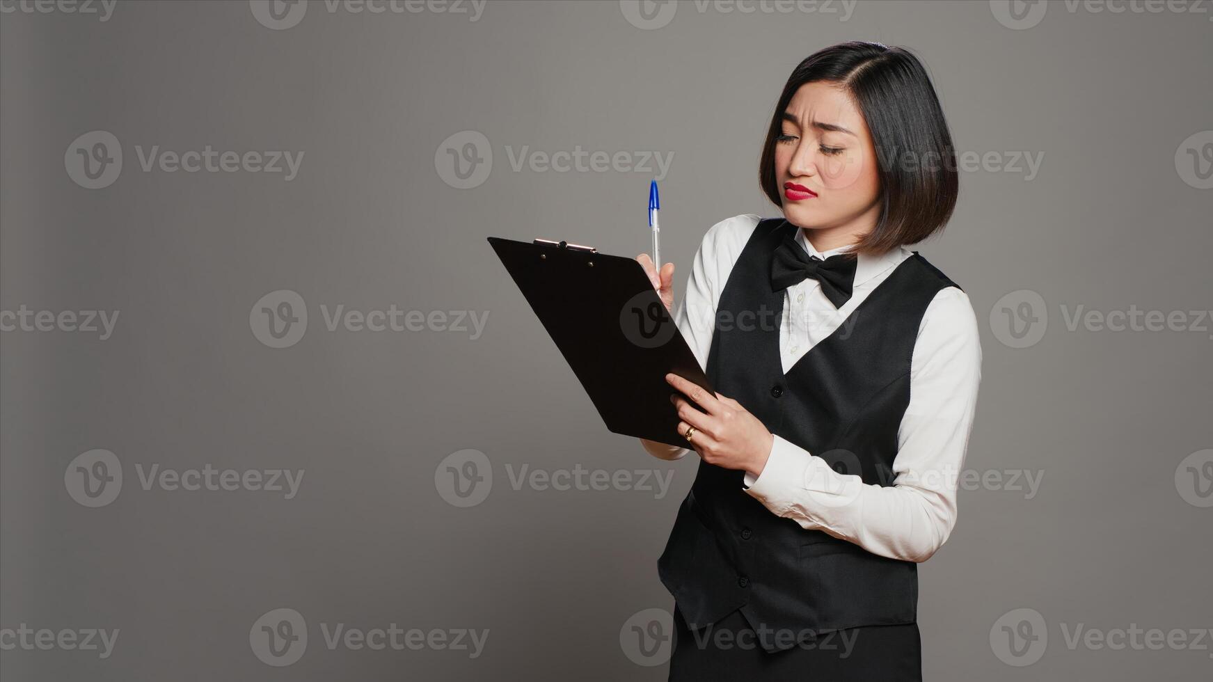 Hotel administrator checking off names of room reservations list, working in hospitality industry to greet clients. Asian receptionist examining registration notes on clipboard files. Camera A. photo