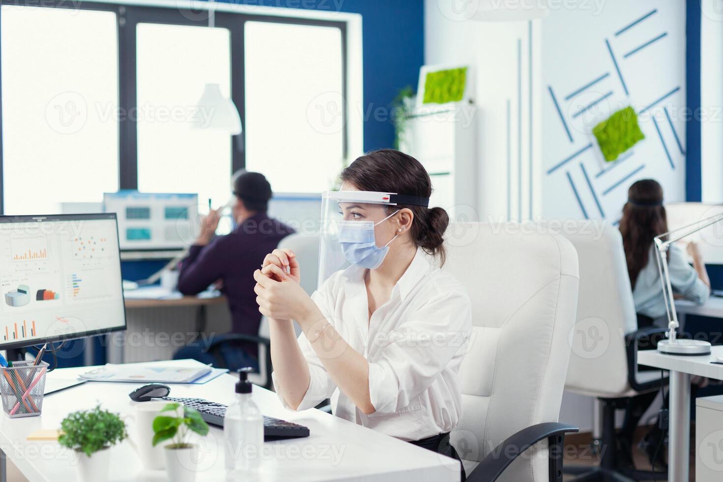 Executive person rubbing hands together after applying sanitizer wearing face mask during global pandemic with covid19. Businesswoman in new normal workplace disinfecting while colleagues working in background. photo