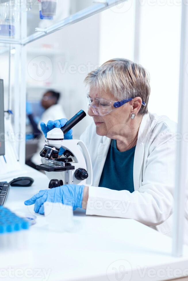 Senior scientist woman with protection glasses doing genetic investigation using microscope with african assistant in the background.. Elderly researcher carrying out scientific research in a sterile lab using a modern technology. photo