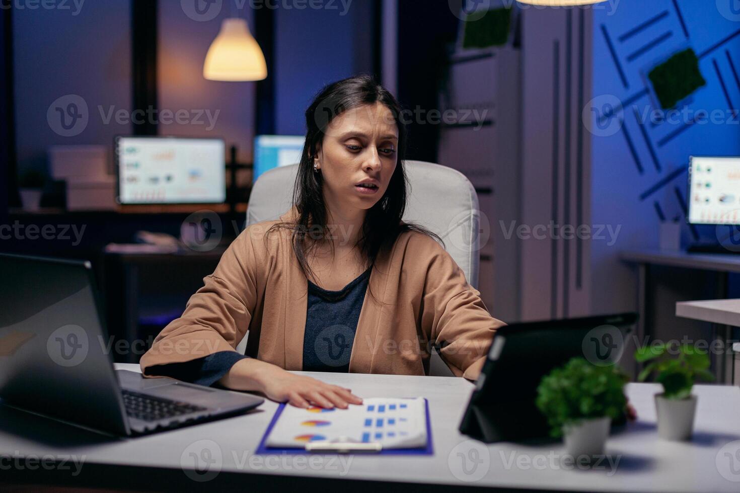 Entrepreneur using tablet pc while working from dark office working on deadline. Business woman working overtime at the office to finish a corporate job using tablet pc. photo