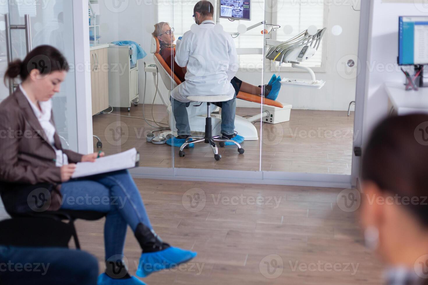 Senior woman sitting on chair in dental office listening dentist talking about diagnosis for teeth cavity looking at digital x-ray radiography. Patients filling form in waiting area. photo