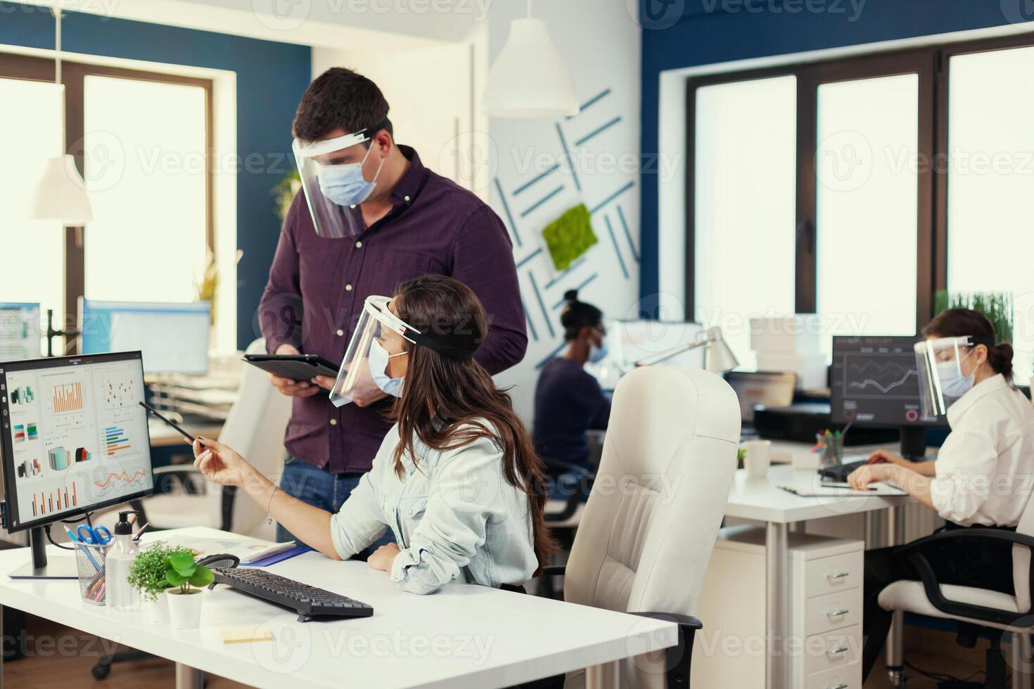 Company analysts helping each other to finish a project at workplace wearing face mask as safety precaution for covid19. Multiethnic team in new normal financial office in corporate company typing on computer, taking notes on tablet. photo