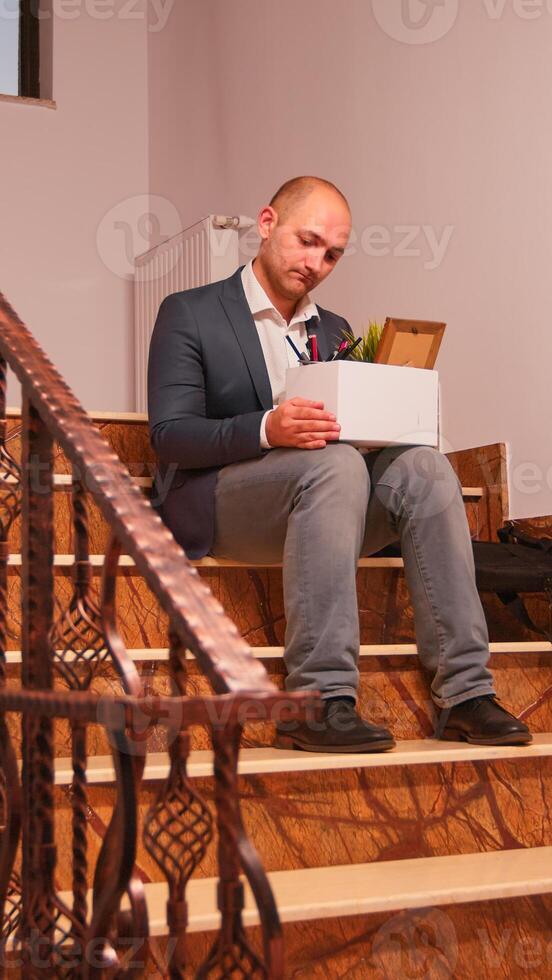 Upset business corporate employee sitting on stairs holding box of personal stuff, getting fired from company. Group of professional successful businesspeople working in modern financial building. photo