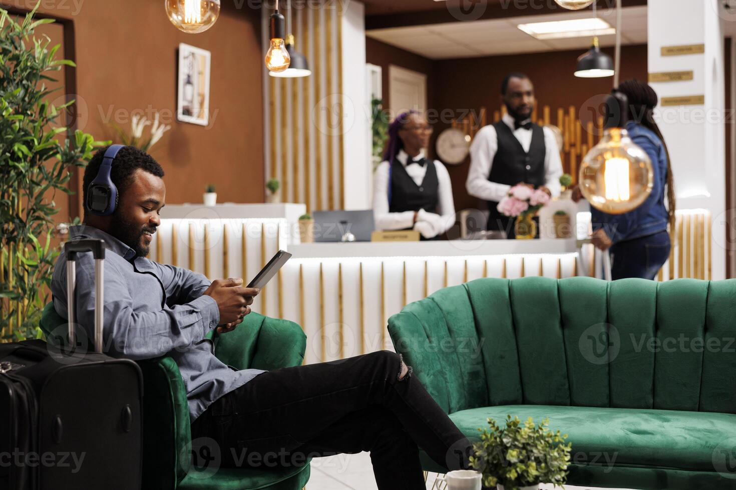 Happy smiling black guy sitting with luggage in hotel lobby in wireless headphones using digital tablet. Young African American man traveler watching movie while waiting for check-in photo