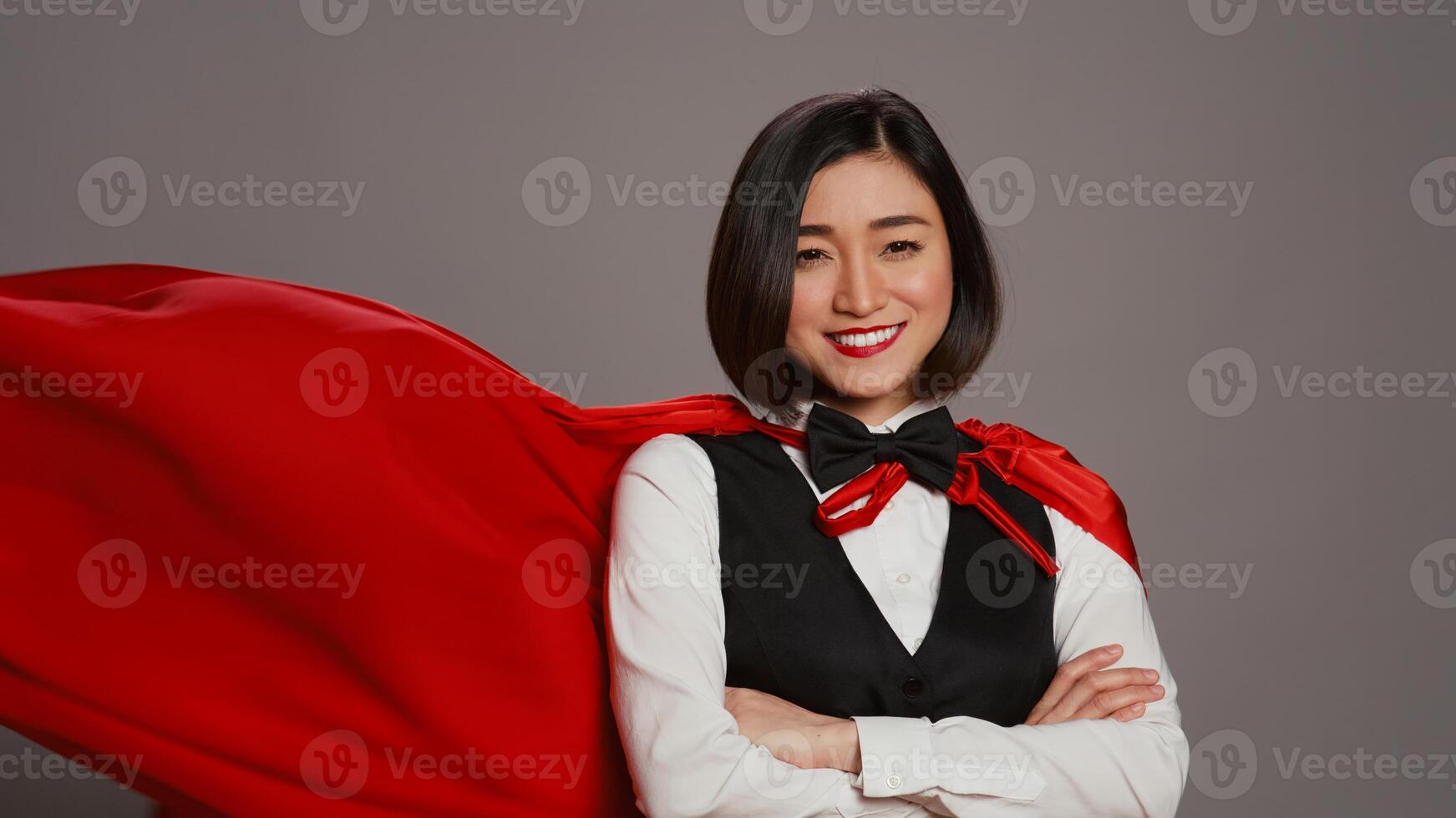 Asian receptionist posing with red superhero cape on camera, feeling confident and powerful while she wears a cloak as a hero. Hotel concierge operator standing with arms crossed. Camera B. photo
