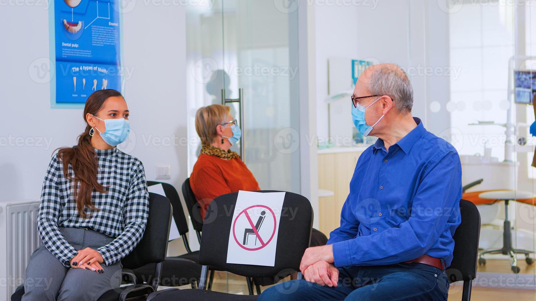 Patients with protection mask talking sitting on chairs keeping social distance in stomatological clinic, waiting for doctor during coronavirus. Concept of new normal dentist visit in covid19 outbreak photo