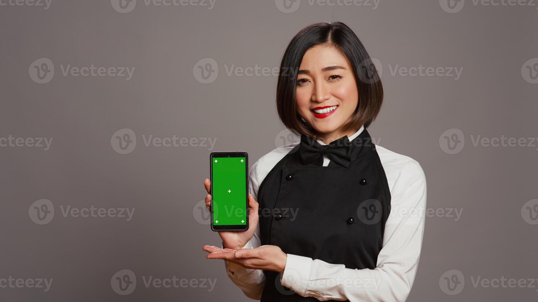 Restaurant hostess presenting smartphone with greenscreen, showing isolated display with chromakey layout in studio. Asian waitress with apron holding phone with copyspace. Camera B. photo
