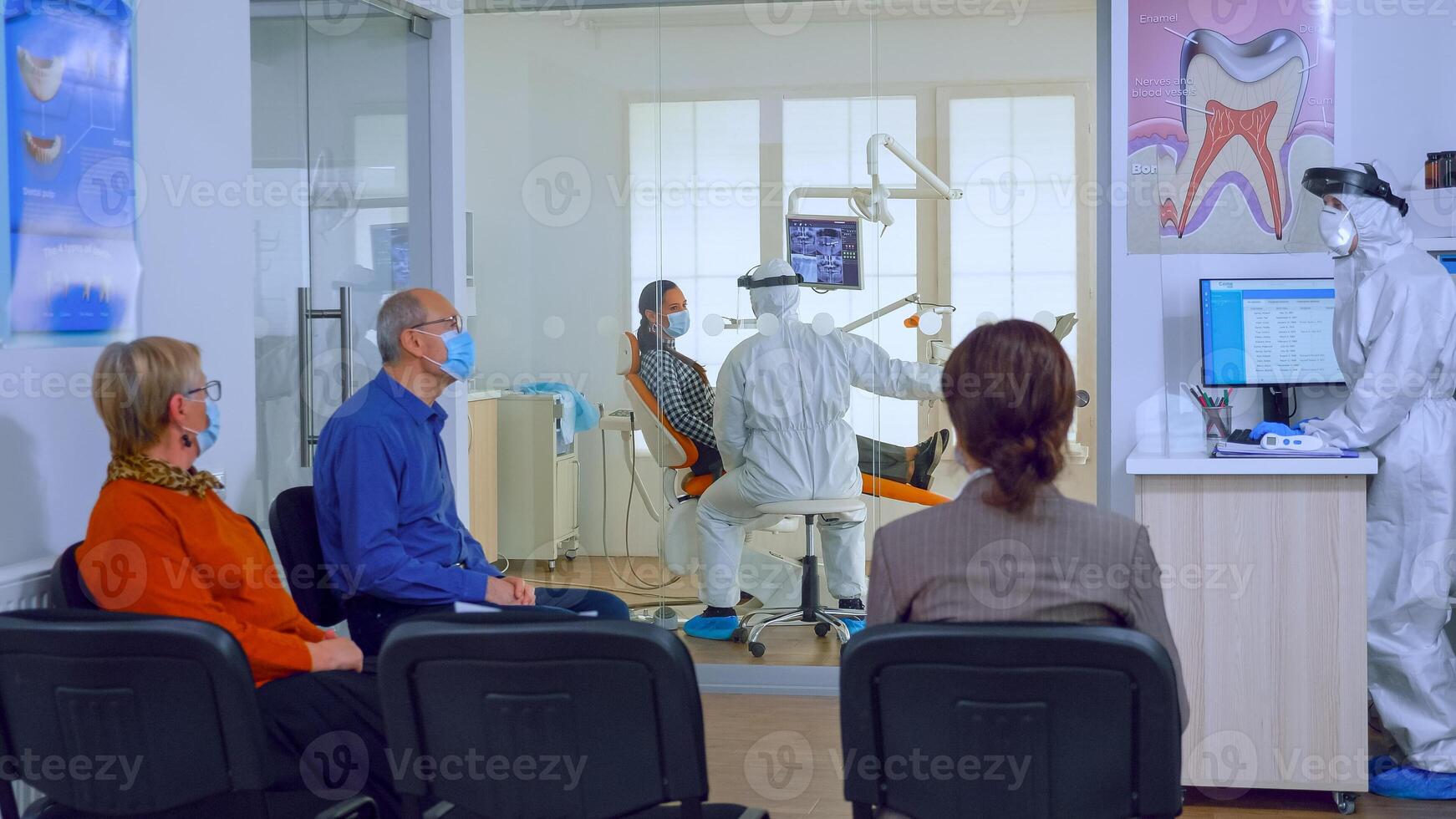 Patients with protection masks waiting doctor in dental clinic reception while stomatologist working in background wearing ppe suit. Concept of new normal dentist visit in coronavirus outbreak. photo