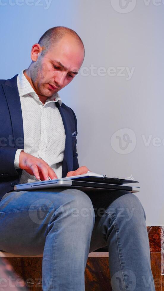 Businessman sitting on stairs in corporate bulding analyzing documents from clipboard overworking in financial company. Office executive checking annual graphs late finishing business deadline project photo