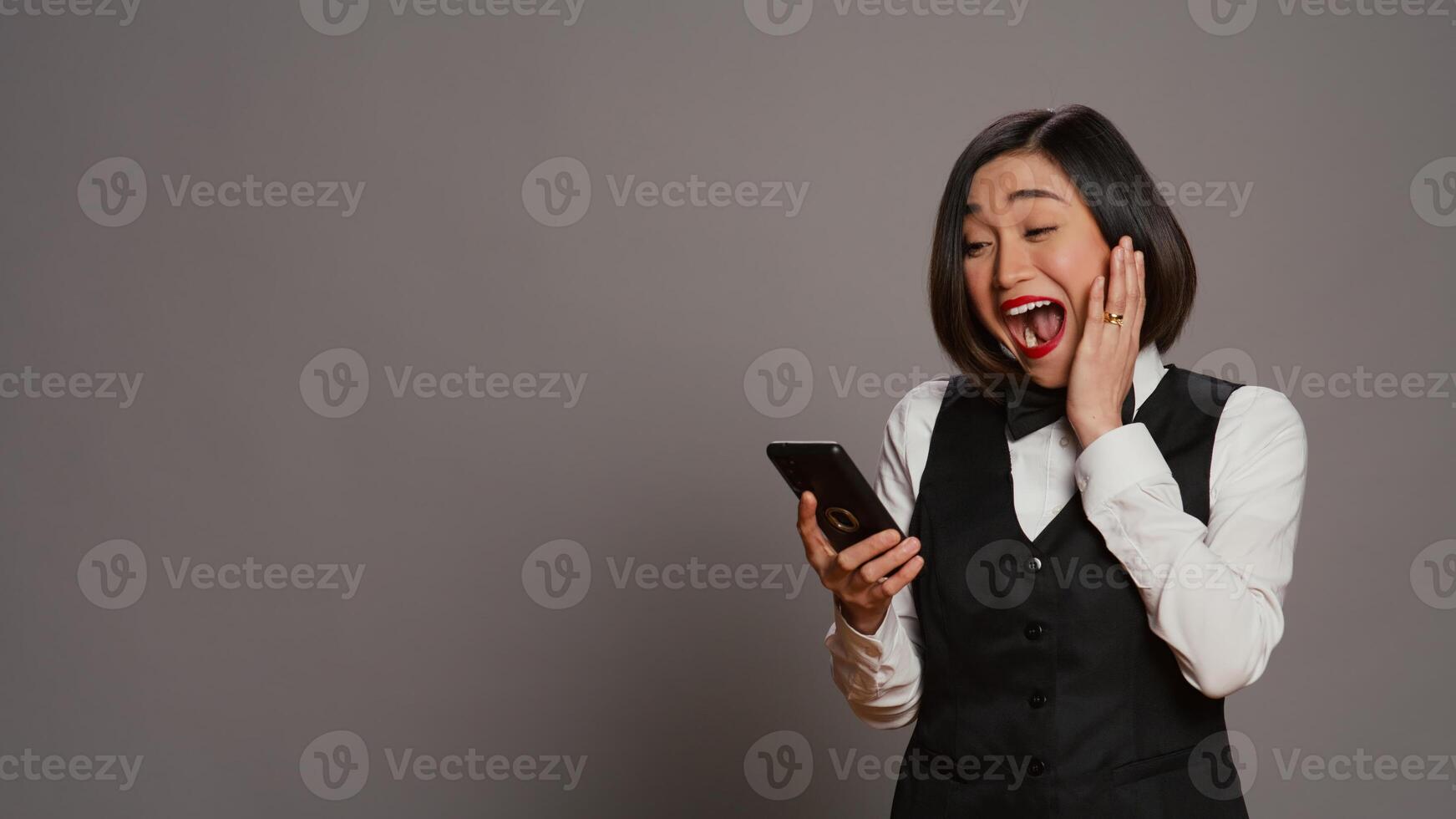 Front desk staff in uniform looks at online information on mobile phone, checking all website reservations over grey background. Asian receptionist using smartphone for bookings. Camera B. photo