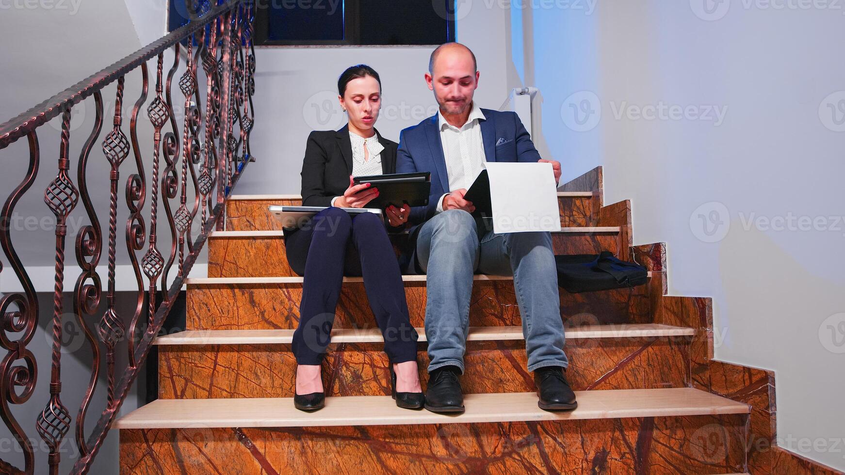 Teamworkers sitting on stairs in corporate building doing overtime during deadline financial project looking on tablet and documents. Entrepreneurs working late together at corporate job. photo