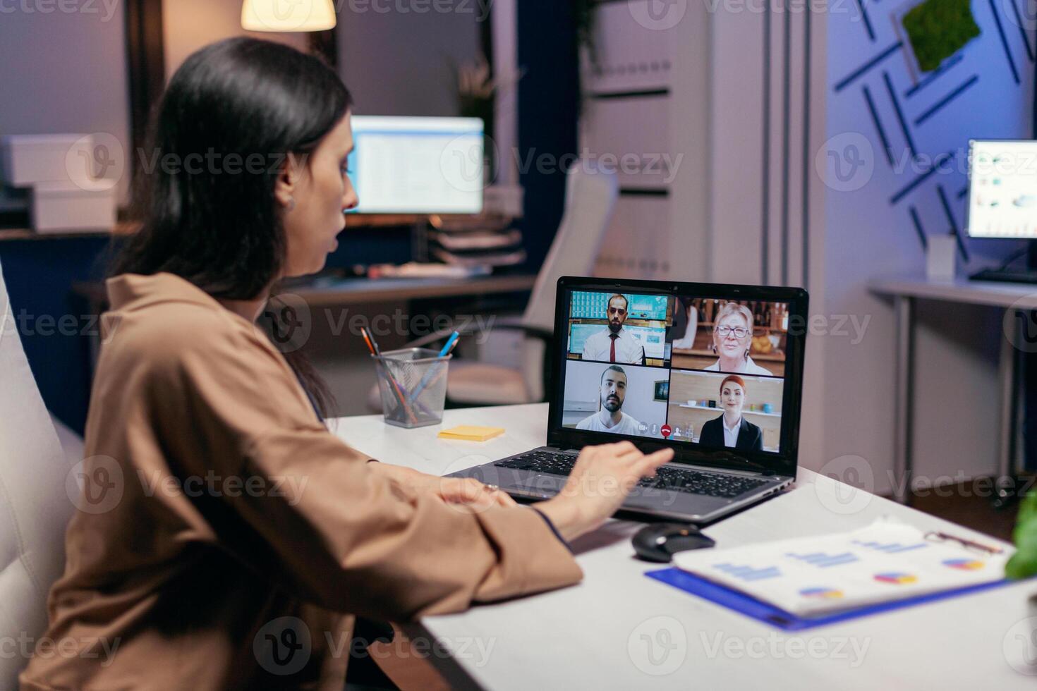 Businesswoman leading a video conference call late night discussing about project deadline. Freelancer working on finance during a video conference with coworkers at night hours in the office. photo