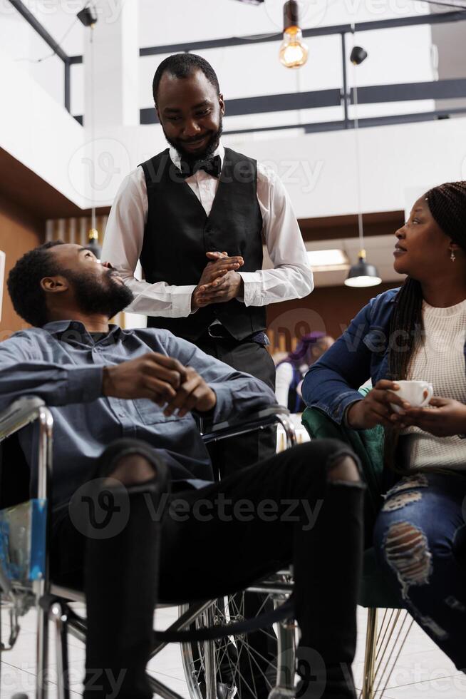 Friendly smiling hotel worker offering assistance to disabled guest on wheelchair, greeting welcoming handicapped man in lobby. Traveler with disabilities asking for help at holiday resort photo