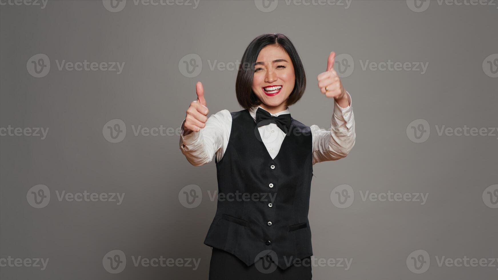 Asian reception staff doing thumbs up symbol on camera, showing approval and positive sign over grey background. Hotel receptionist giving like and okay gesture, showing agreement. Camera A. photo