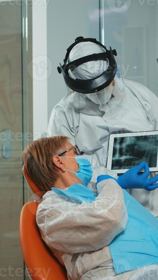 Stomatologist in protective suit reviewing x-ray of tooth with senior patient explaining treatment using tablet in covisd-19 pandemic. Medical team wearing face shield, coverall, mask and gloves. photo