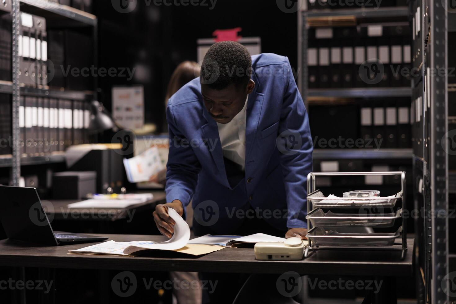 Depository worker reading administrative files, working late at night at management research in storage room. African american bookkeeper analyzing accountancy report in corporate repository photo