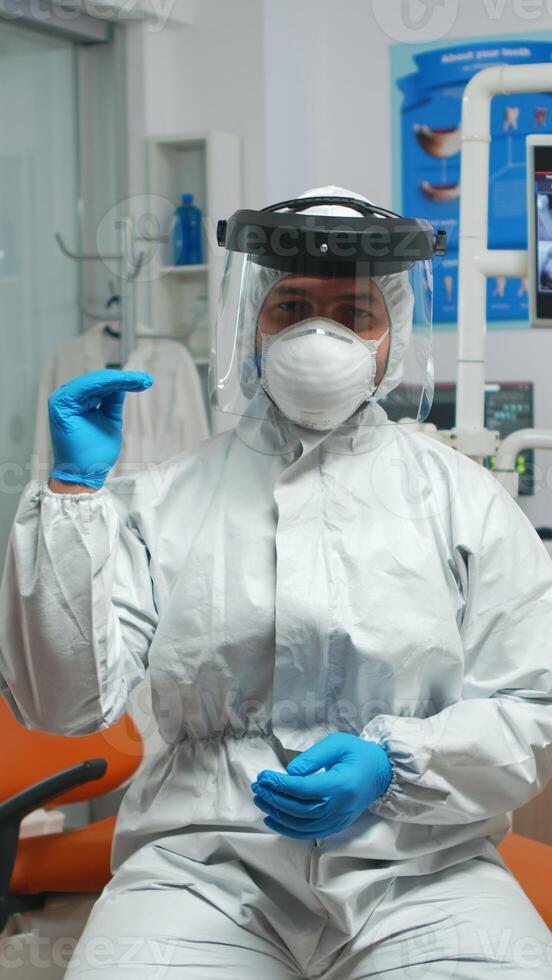 Dentist doctor with coverall looking at camera talking during coronavirus. Orthodontic on video call wearing protection suit, face shield, mask, gloves with assistant in background in pandemic period photo