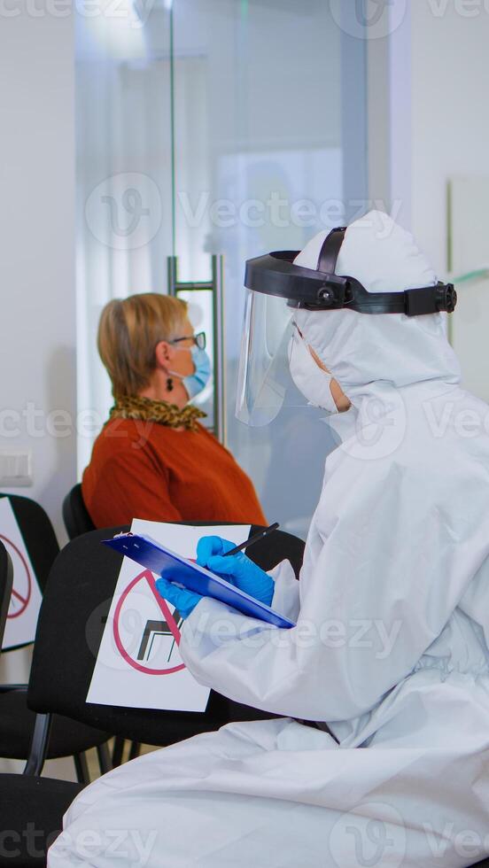 Stomatologist in protective suit reviewing registration form with patient explaining treatment writing on clipboard in covid-19 pandemic. Medical nurse wearing face shield, coverall, mask and gloves. photo