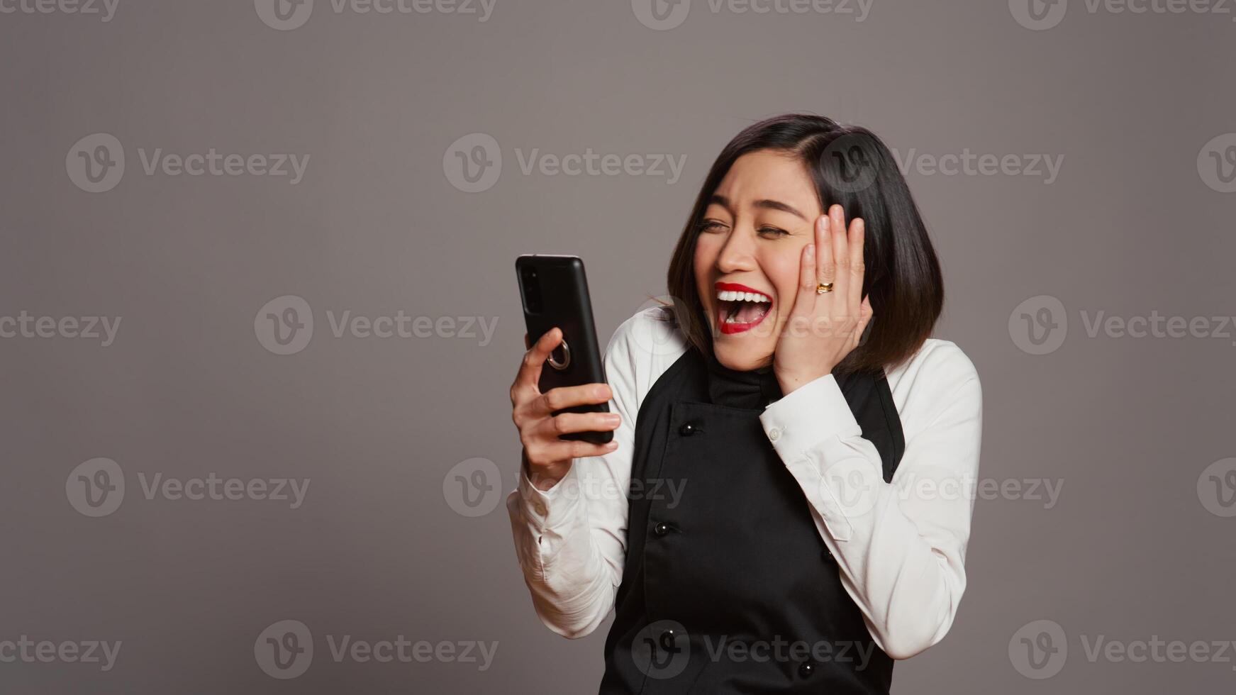 Asian server feeling happy after reading good news on phone, having a lot of clients with reservations for dinner. Waitress with apron using smartphone to see table bookings, enjoys work. Camera B. photo