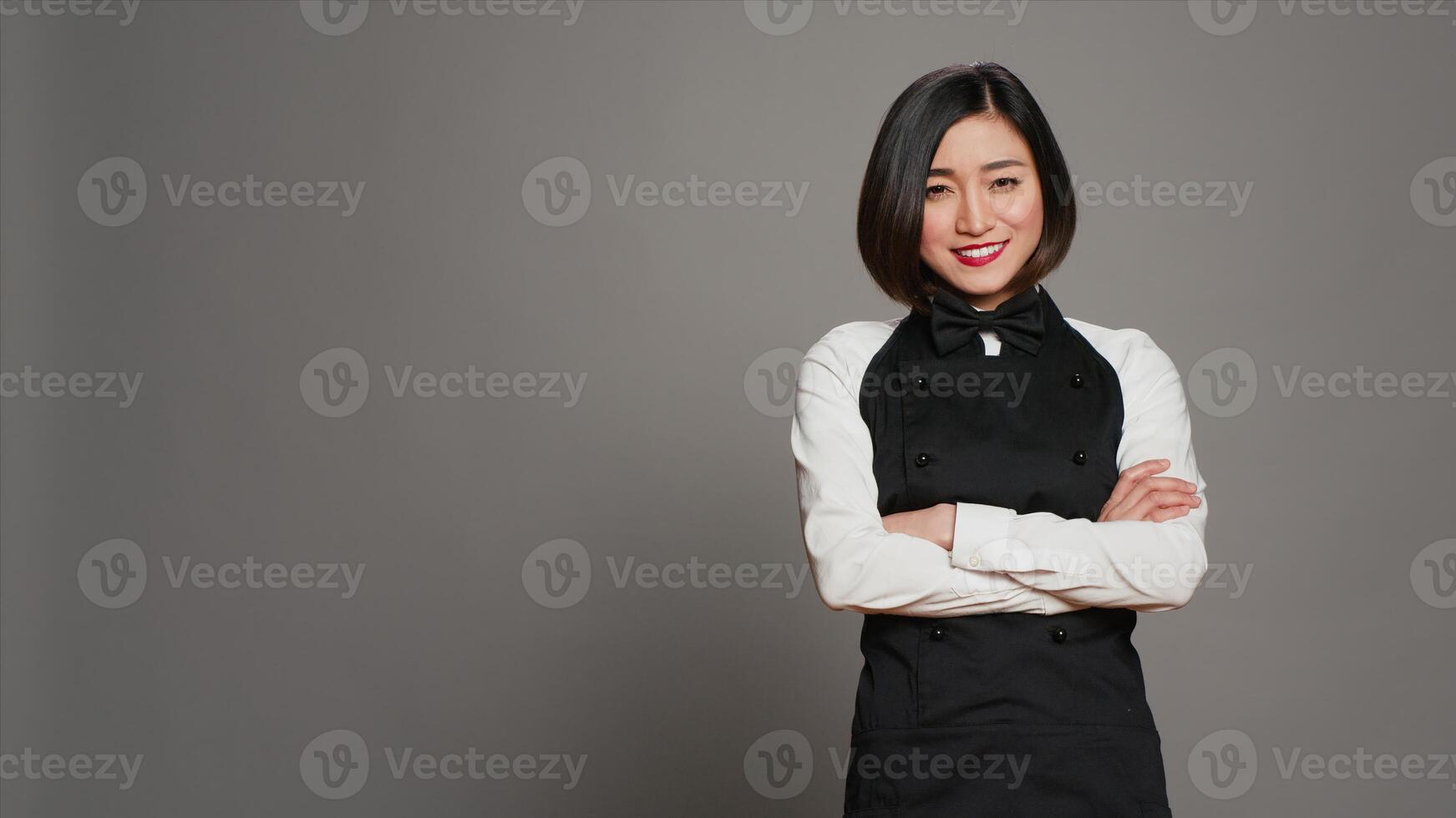 Asian diner employee prepared to serve food at tables, working in hospitality cuisine and catering. Restaurant waitress with apron standing over grey background, gourmet service. Camera A. photo