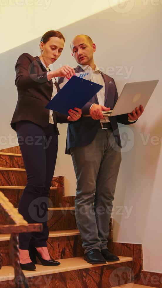 Corporate executive manager holding laptop talking with businesswoman on staircase of business building. Professional successful entrepreneur in team working overtime in modern office photo