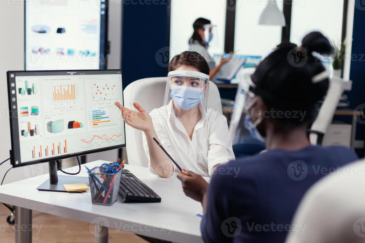 Manager wearing face mask for covid19 explaining financial graph to african employee. Multiethnic team working in company with new normal respecting social distance because of global pandemic with coronavirus. photo