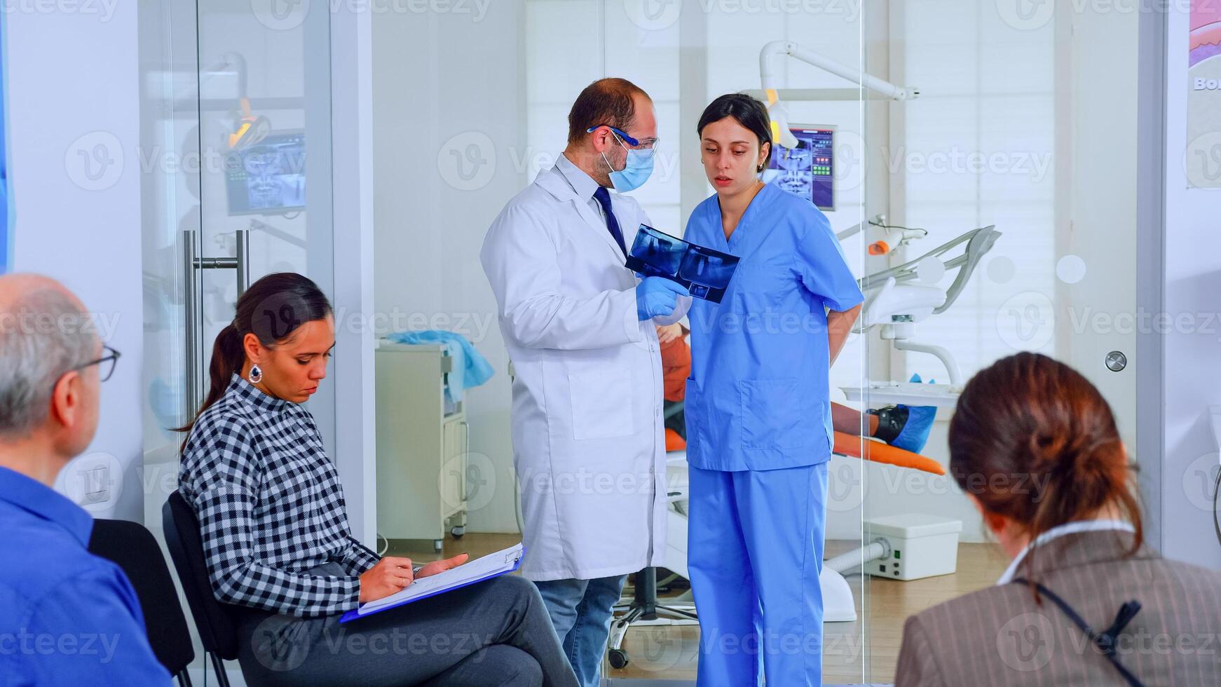 Dentist showing teeth x-ray reviewing it with nusre. Doctor and assistant working in modern crowded stomatological clinic, patients sitting on chairs in reception filling in dental forms and waiting photo