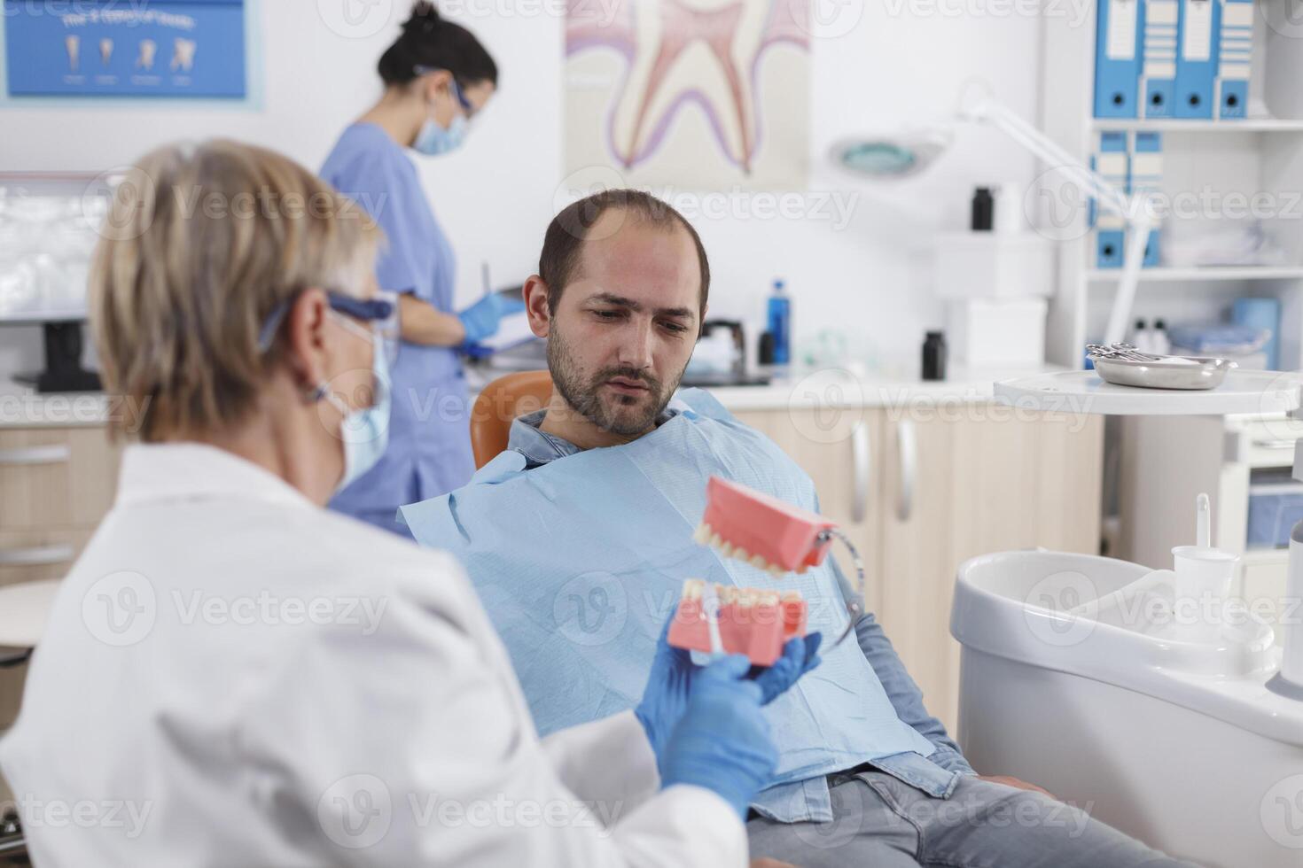 Senior woman doctor showing artificial jaw model to patient with toothache discussing carier treatment during stomatology consultation in dentistry office room. Stomatologist explaining oral hygiene photo