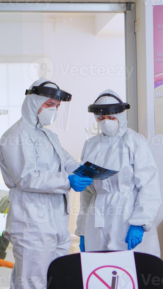 Dental doctors with protection suit analysing teeth x-ray in reception while patient waiting on chair, respecting social distance. Concept of new normal dentist visit in coronavirus outbreak. photo