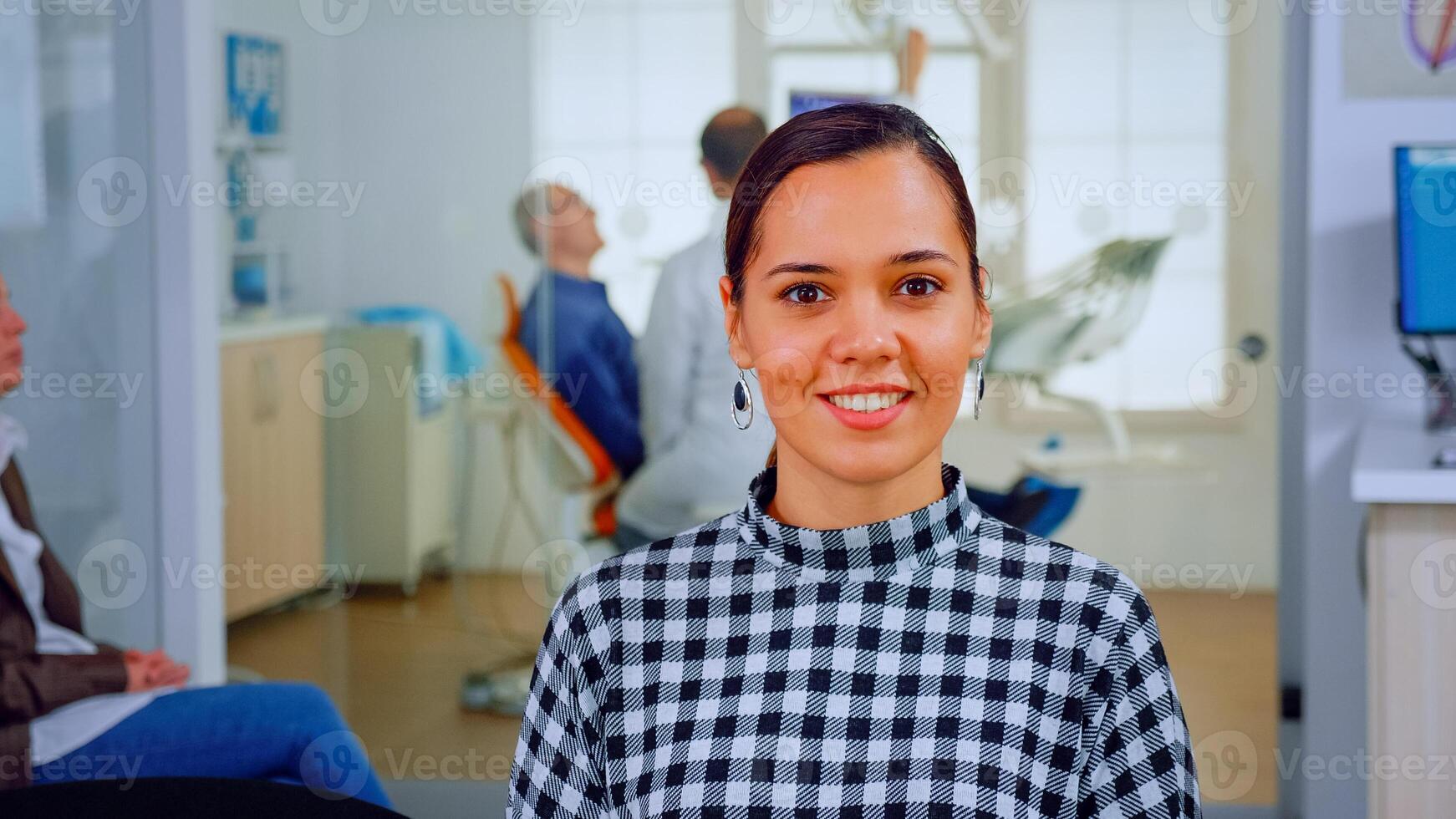 Portrait of smiling woman patient looking on webcam sitting on chair in waiting room of stomatological clinic. Stomatologist assistant typing on pc in dental office while doctor working in background. photo