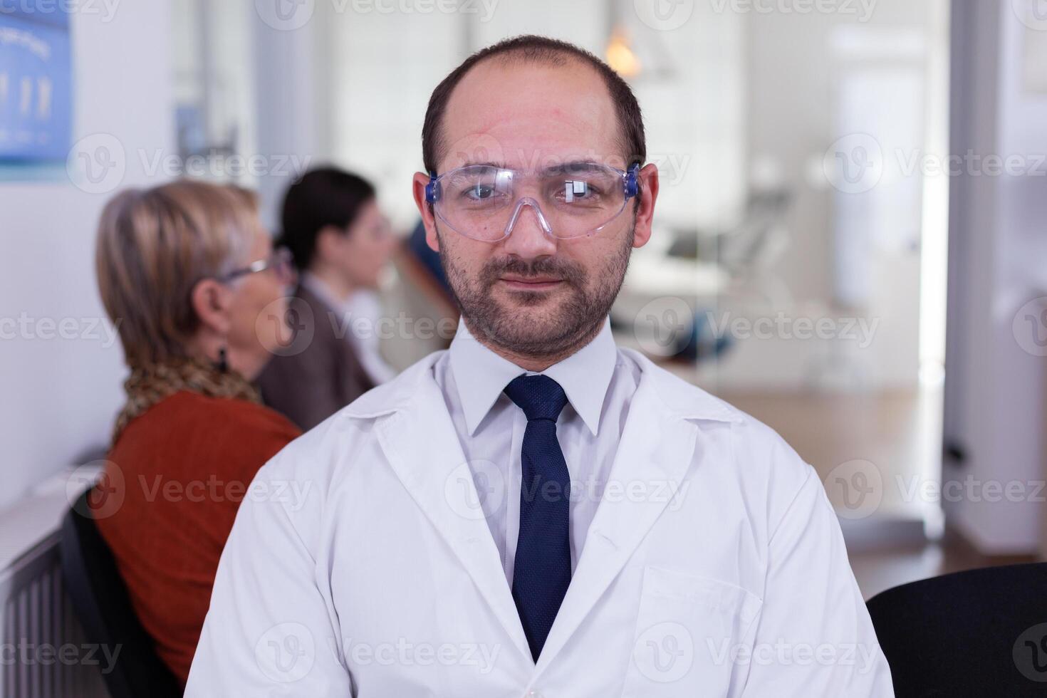 Portrait of stomatologist smiling at camera being in dental office while patients waiting him in background. Dentistry doctor looking on webcam sitting on chair in stomatological clinic. photo