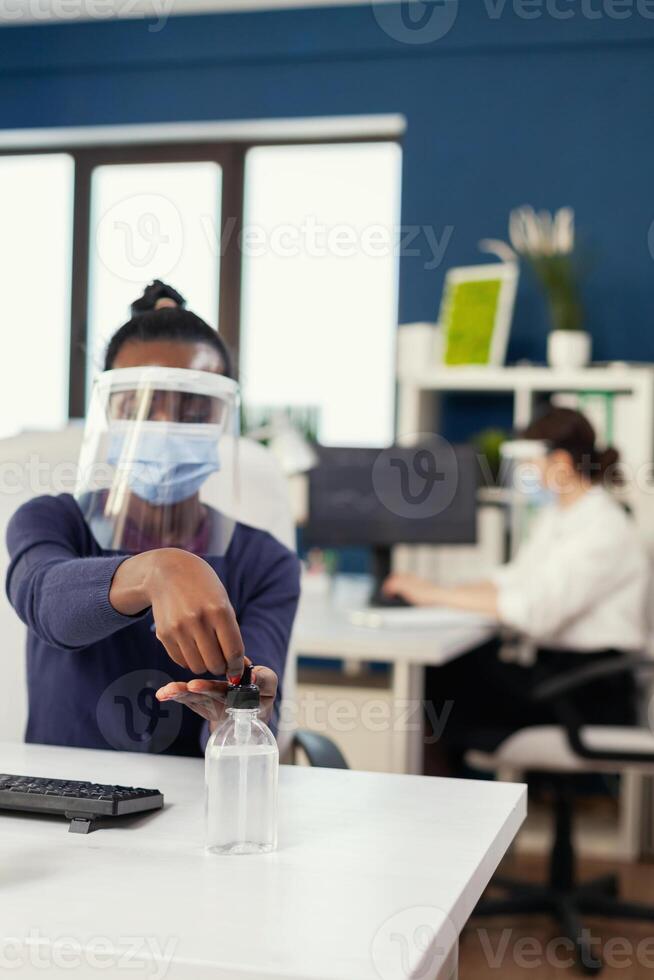 africano mujer en sociedad oficina aplicando mano desinfectante vistiendo cara máscara durante coronavirus. mujer de negocios en nuevo normal lugar de trabajo desinfectar mientras colegas trabajando en antecedentes. foto