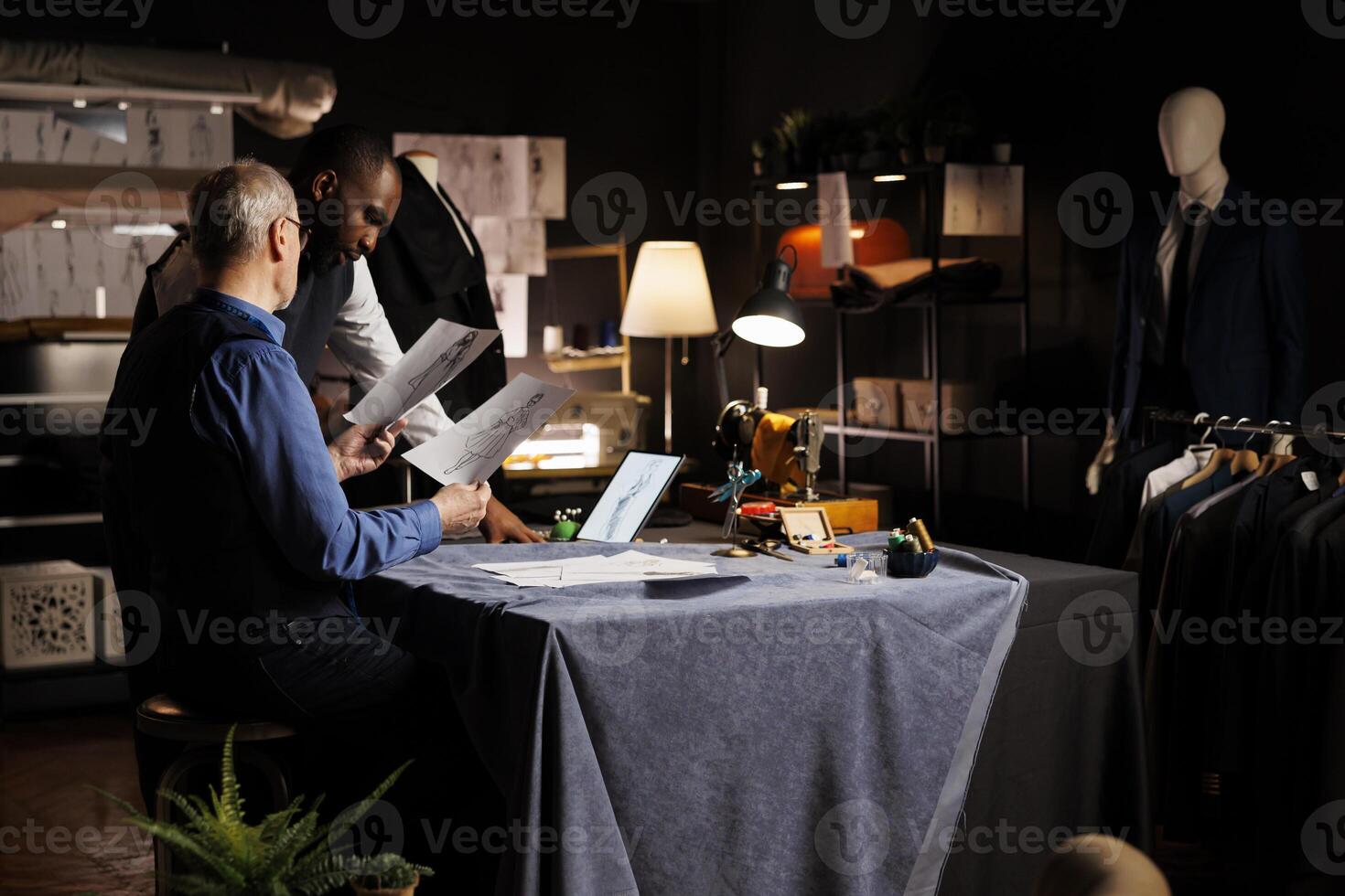 Skilled dressmakers working on upcoming refined modern fashion design collection, comparing dress sketches. Masterful experienced creative gentlemen creating fashionable bespoke attire in dark atelier photo