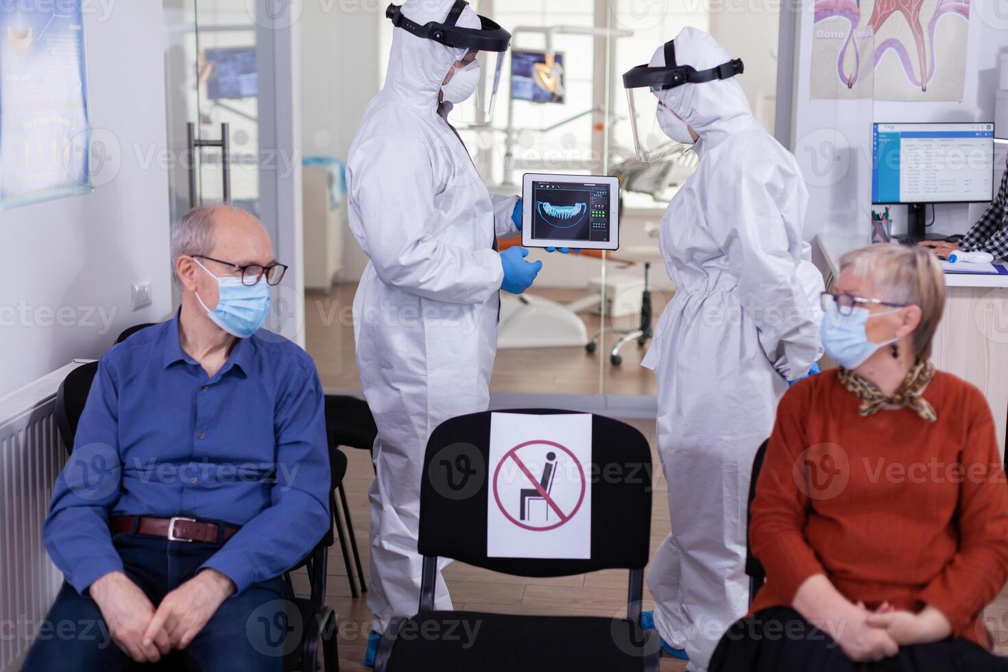 Man discussing with nurse in dental reception wearing protection suit against coronavirus, elderly patients waiting in reception keeping distance. Concept of new normal dentist visit in outbreak. photo