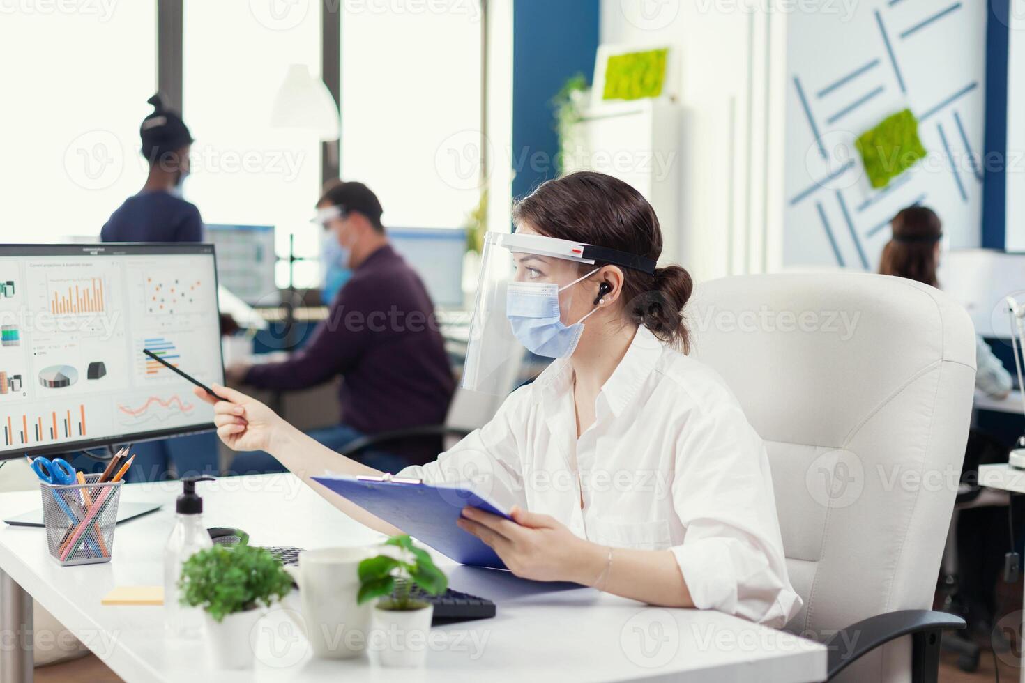 Businesswoman looking at financial stats wearing face mask during covid-19. Employees with visors working in corporate company workspace respecting social distance analysing data and graphs. photo
