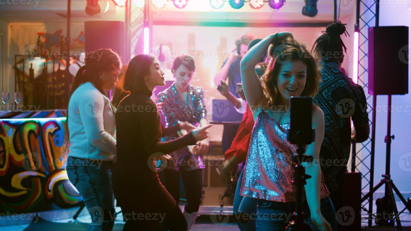 Happy girl recording vlog for internet, using mobile phone at discotheque on dance floor. Female influencer filming cool video of people dancing together and jumping, disco event. Handheld shot. photo