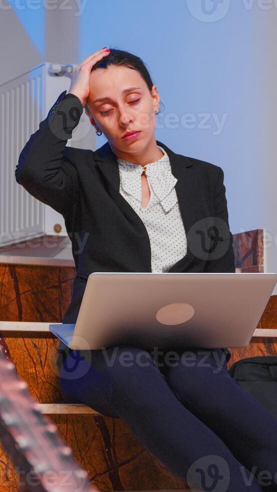 Overworked tired businesswoman with headache working on difficult project deadline using laptop sitting on staircase of business building. Serious entrepreneur being on corporate job late at night. photo