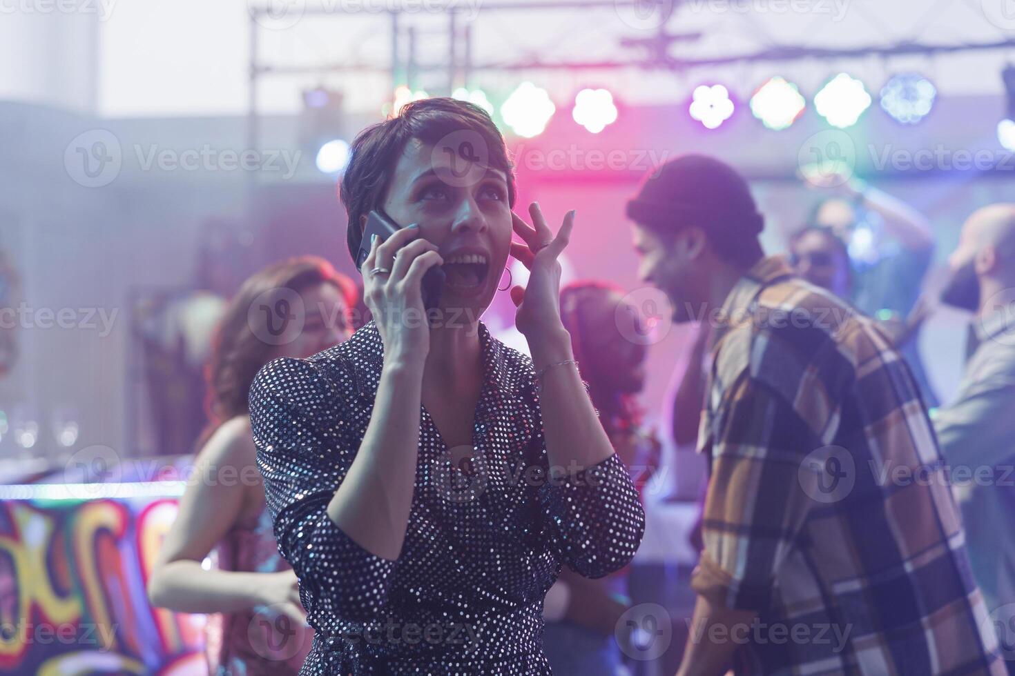 Young woman shouting and covering ear while trying to talk on smartphone in nightclub with loud music. Clubber girl answering mobile phone call while partying in club gathering photo