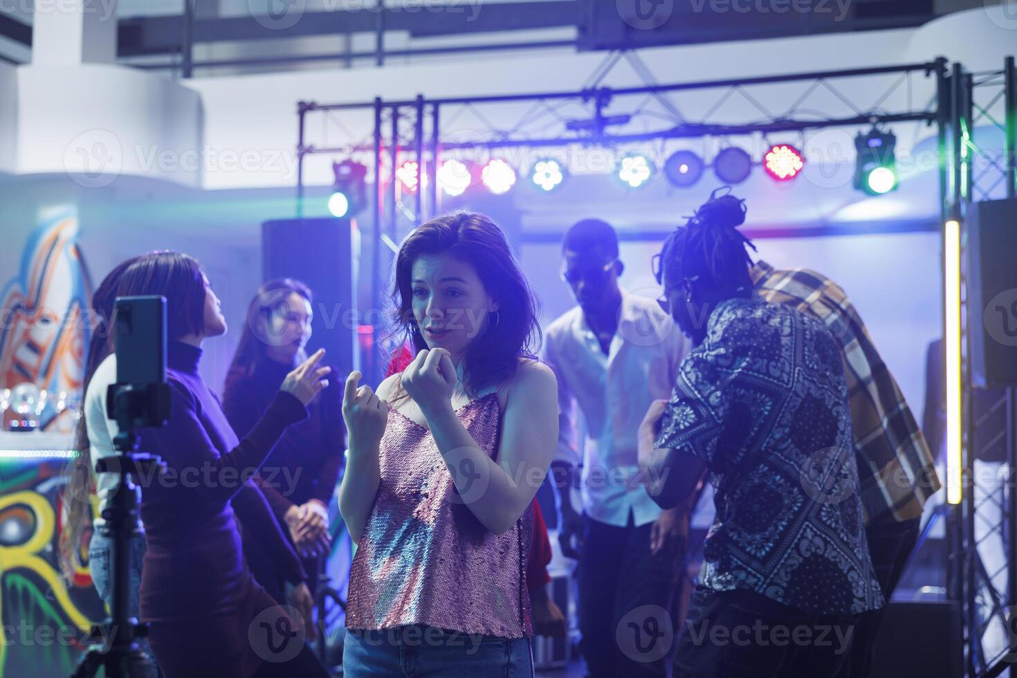 Young woman speaking on smartphone camera while recording video at nightclub party. Clubber girl having fun at club gathering and broadcasting online using mobile phone on tripod photo