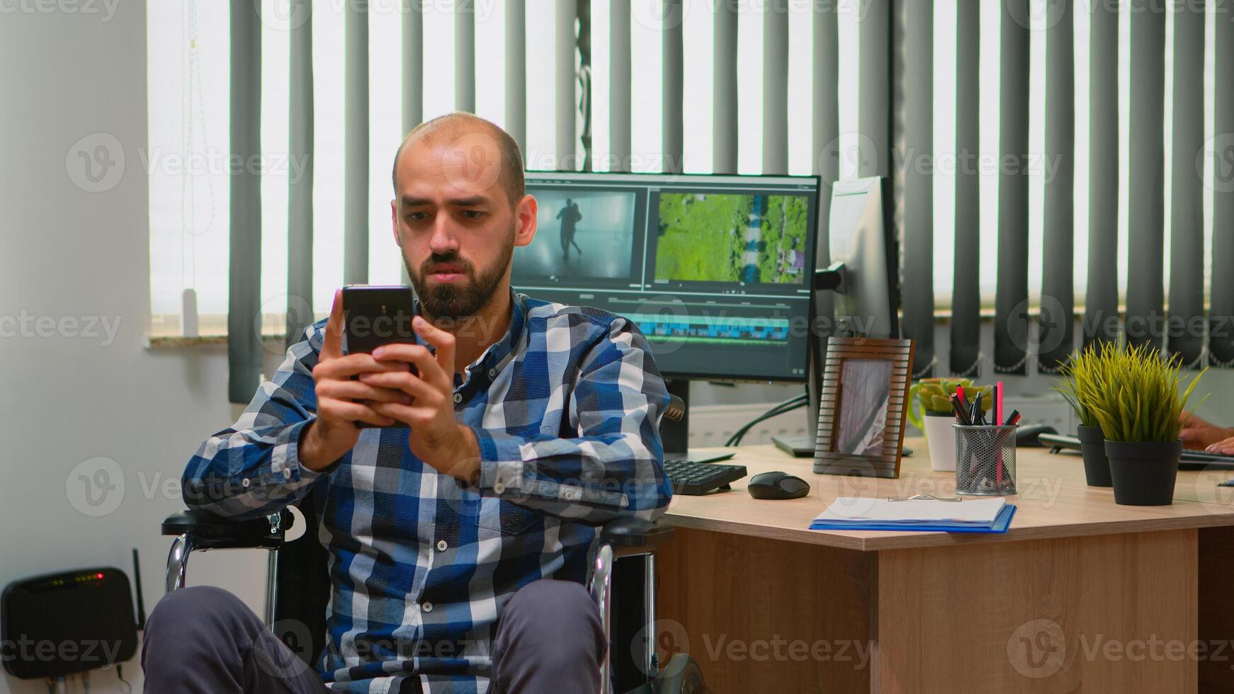 Freelancer photo designer with disability in wheelchair texting on smartphone taking break from editing video project creating content in modern company office. Blogger working from photo studio.