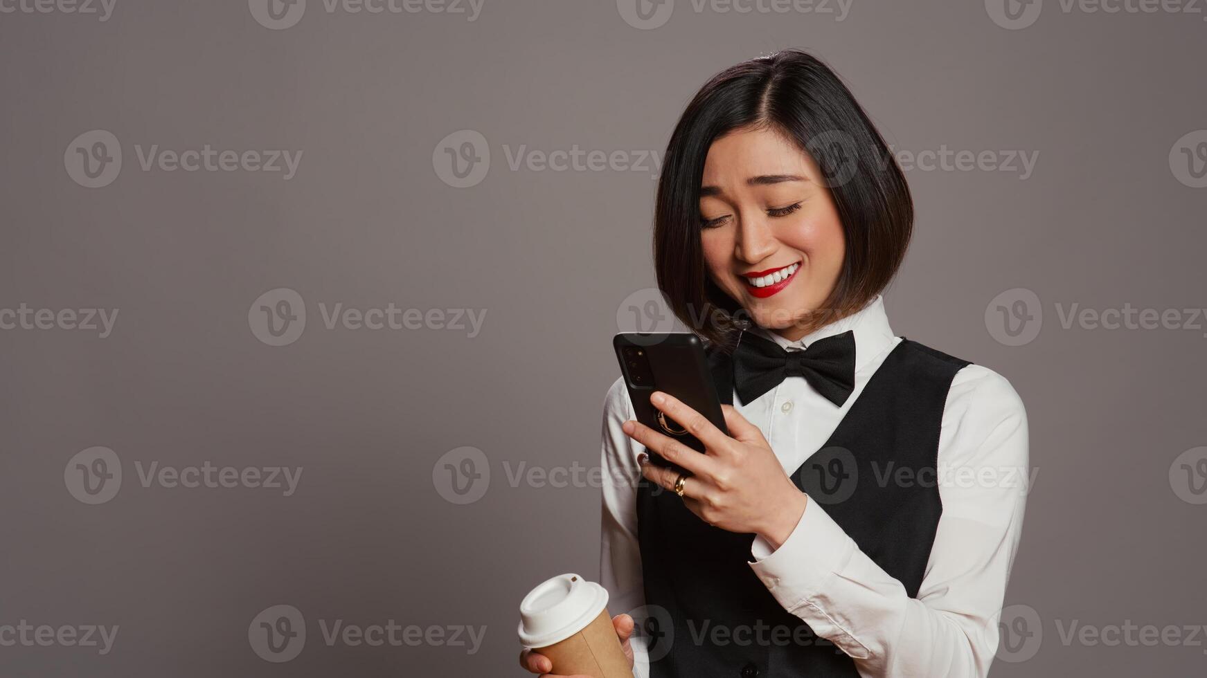 conserje personal Bebiendo taza de café en estudio, hojeada teléfono inteligente social medios de comunicación aplicaciones asiático recepcionista disfrutando cafeína refresco mientras ella usos móvil teléfono. cámara b. foto