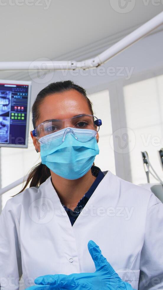 médico medición mujer temperatura antes de dental examen, paciente punto de vista dentista y enfermero trabajando en moderno ortodoncia oficina, escritura y examinando persona vistiendo proteccion máscara foto